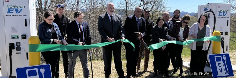 Officials cut a green ribbon in front of two electric vehicle charging stations with a blue sky and trees in the background.