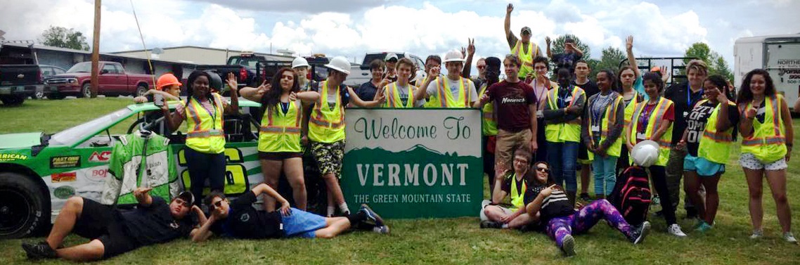 Over 20 Youth with reflective vests pose in front of parking lot with "Welcome to Vermont - The Green Mountain State" sign